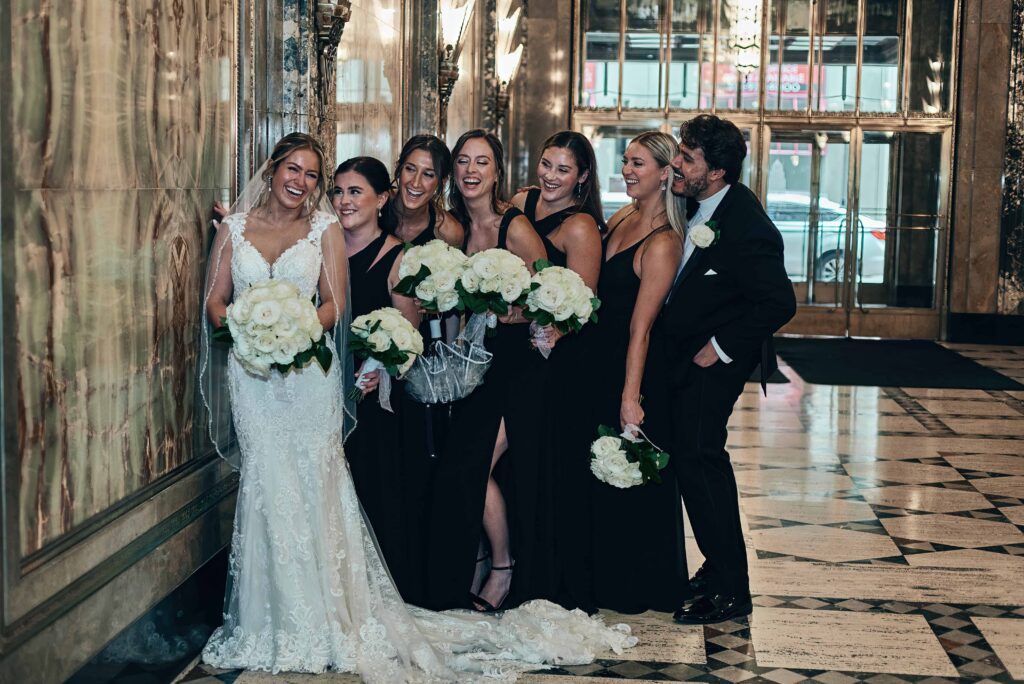 A bride in a white lace dress holding a bouquet of white roses leans agains a marble wall with her five bridesmaids and one bridesman stand in a line next to her looking at her and smiling