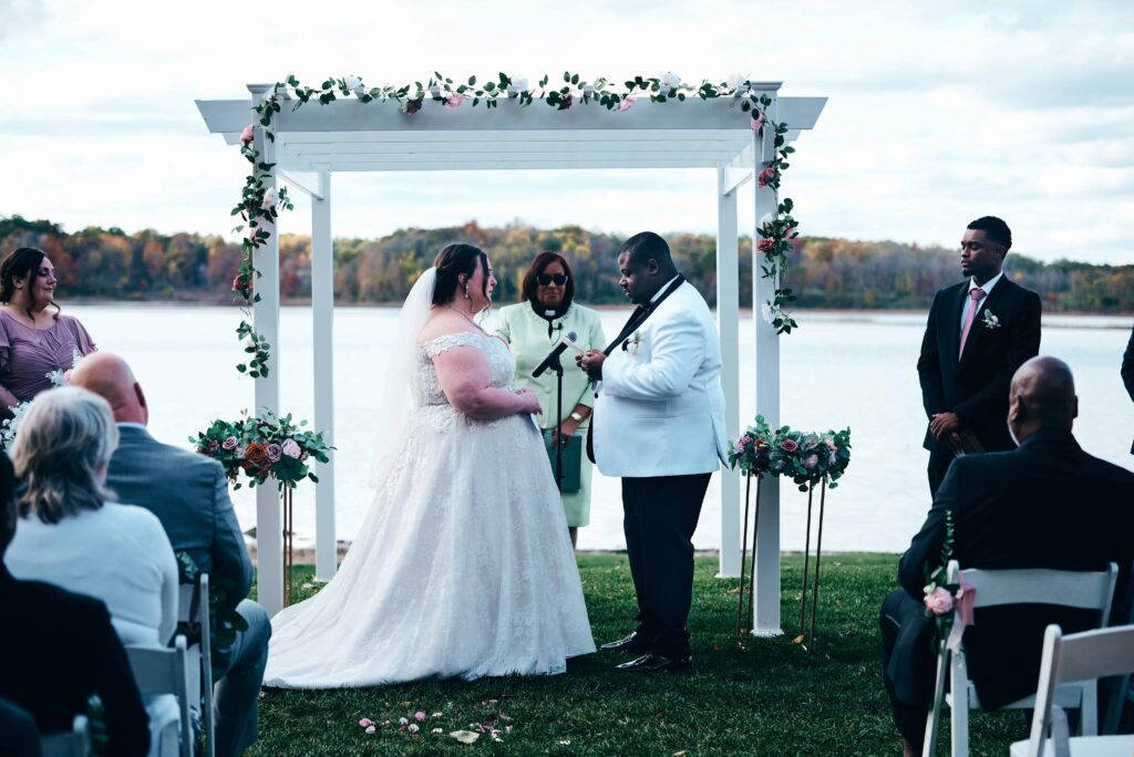 A bride in a white, lace wedding dress and a groom in a white suit exchange vows during a lakeside ceremony with fall leaves lining the opposite shoreline