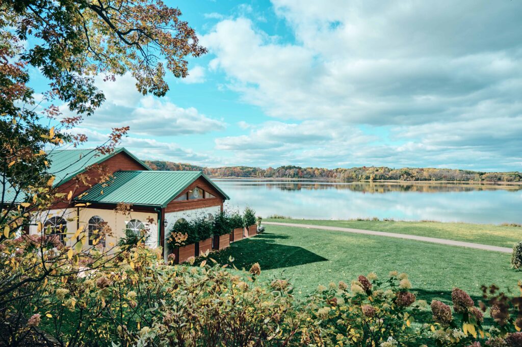 A landscape of blue skies, a lake reflecting that blue sky, a line of fall colors on the opposite shoreline, and a pavilion sitting on a green lawn surrounded by trees