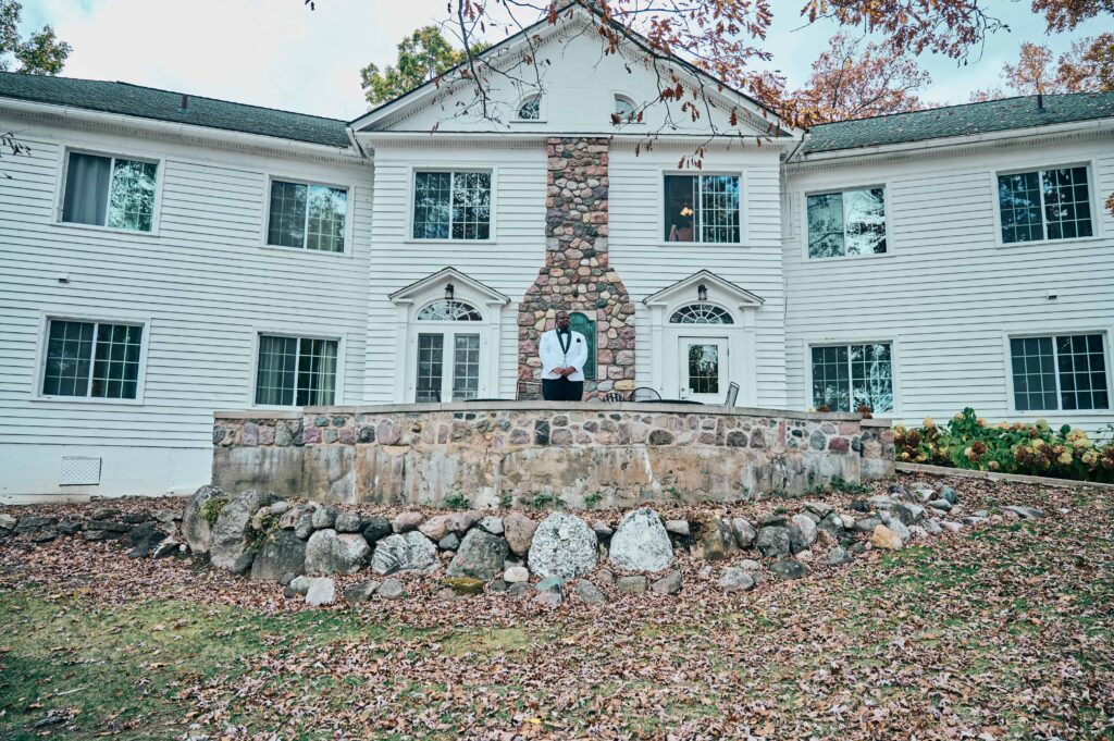 A groom in a white suit stands, hands clasped together, in front of a white building with a large stone fireplace and stone wall