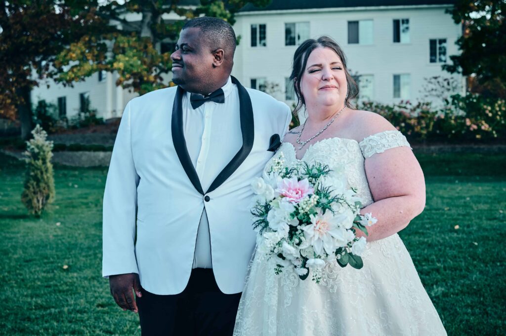 A bride in a white, lace dress with drop cap sleeves and a groom in a white suit with a black bowtie stand close to each other and looking opposite direction. They stand in front of a green lawn and a large white building behind them surrounded by trees.