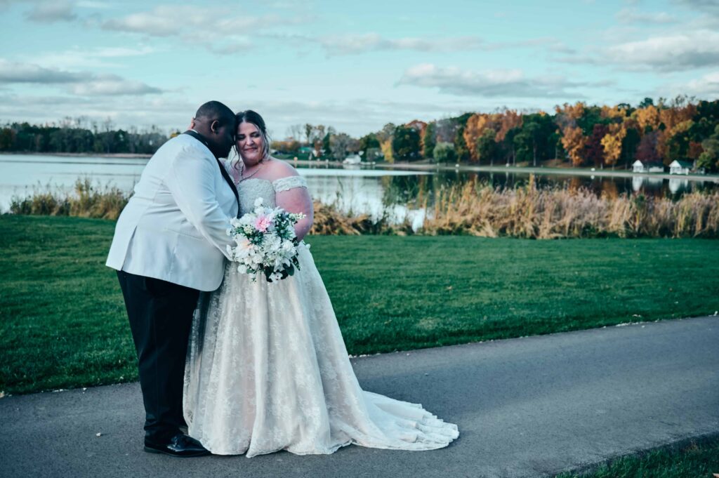 A groom in a white suit nuzzles a bride in a white, lace dress holding a white, pink, and green bouquet. They stand on a path with a green law, a lake, and fall colored trees behind them