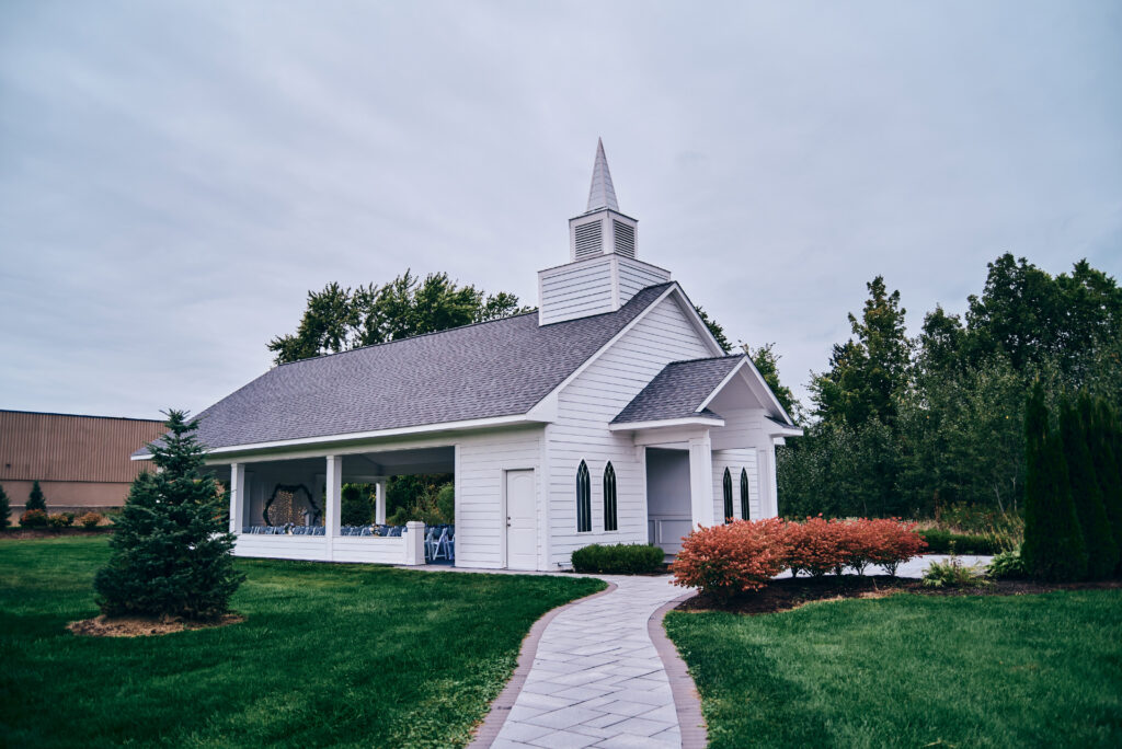 A white wedding chapel with open-air sides. This white chapel is surrounded by green grass and trees and some orange bushes in the front.
