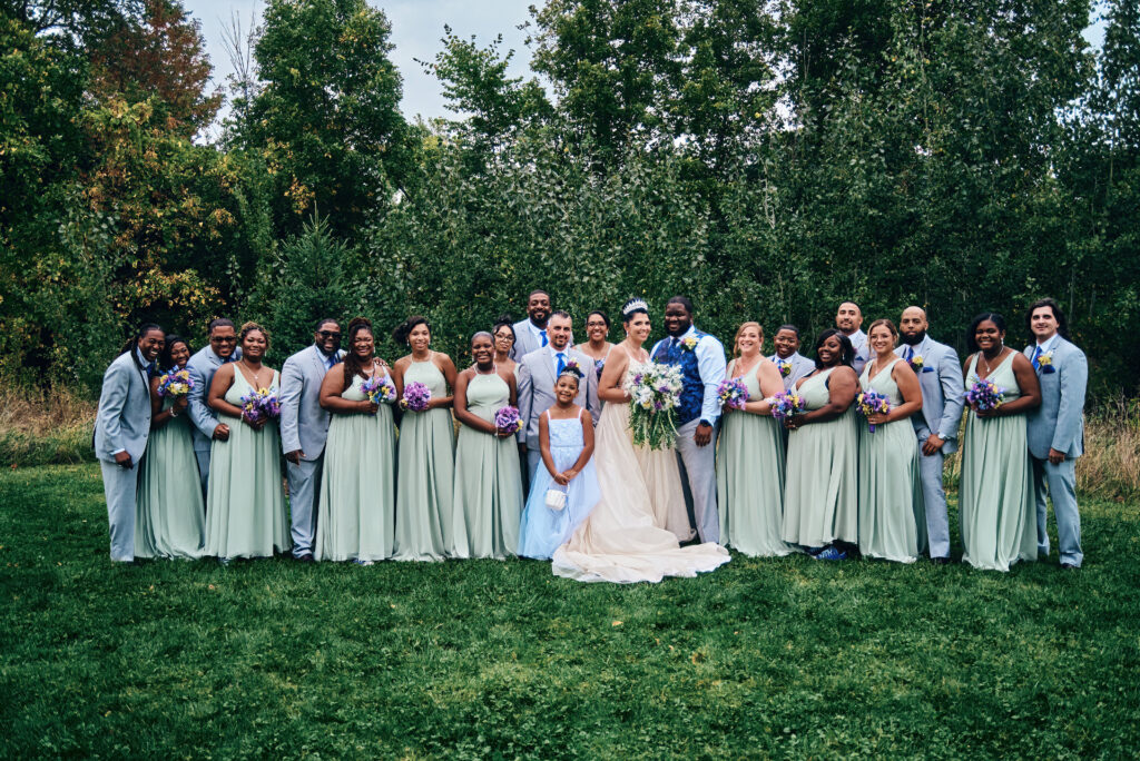 A large wedding party of twenty one people stand in a line surrounding the bride and groom looking at the camera. They are all surrounded by green trees and grass