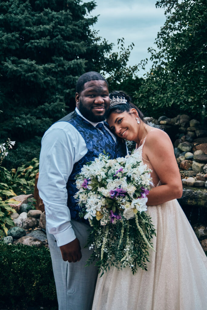 A bride and groom stand together looking at the camera. The bride is leaning her head on the groom's shoulder while she holds her bouquet of pink and purple flowers.