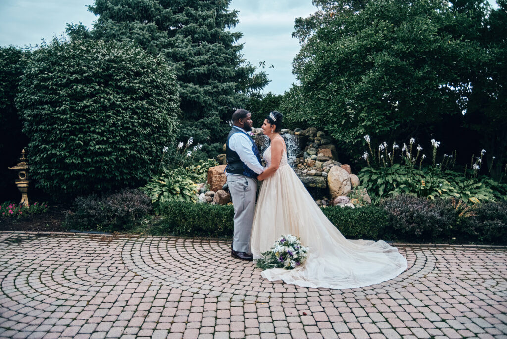 A bride and groom in their wedding attire face each other, holding hands, in front of a small man-made waterfall and standing on a stone-paved walkway. They are surrounded by green trees and plants
