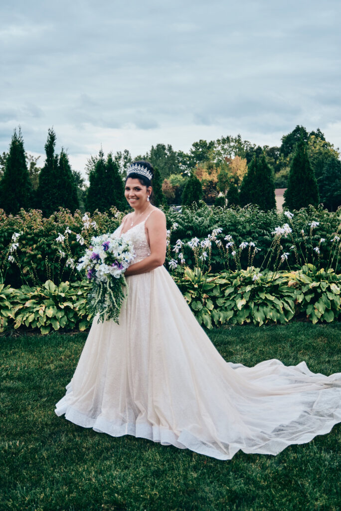 A brunette bride stands in her blush colored sparkly wedding dress in the middle of a garden landscape with hostas and other plants around her. She is holding a bouquet of white flowers and greenery and wears a large, jeweled crown on top of her head.