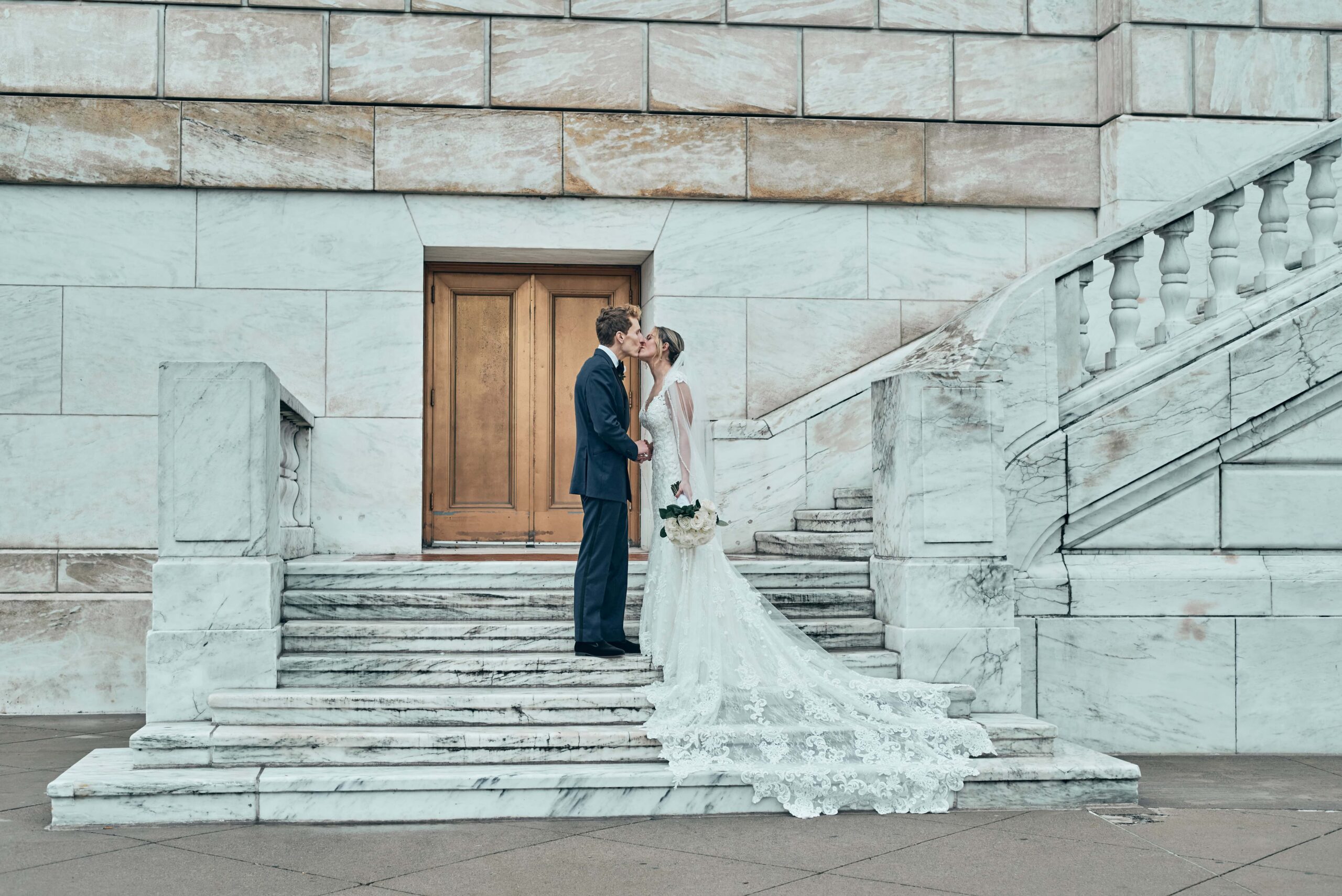 A bride in a long, lace white gown and holding a bouquet of white roses down at her side kisses the groom in a black tux. They stand on white marble stairs with a wall of white marble blocks behind them and a light, brown wooden door.