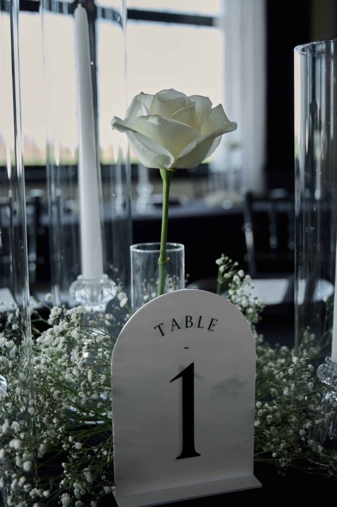A white table number sign with "Table 1" on it stands on a black table cloth in front of white baby's breath and a single white rose.