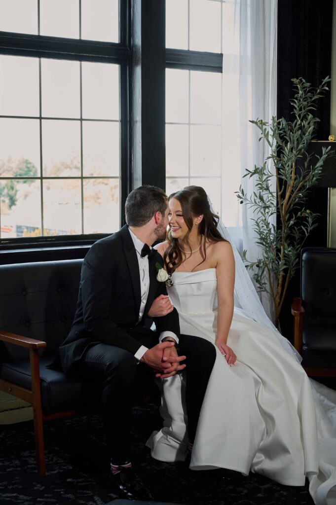 A bride and groom sit on a dark leather couch facing each other, faces close. The bride has her right arm around the groom's left arm while the groom has his hands folded in his lap.